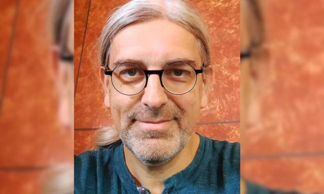 Porträtaufnahme eines Mannes. Er ist Mitte Vierzig und trägt eine Brille, sowie schulterlange weiße Haare.