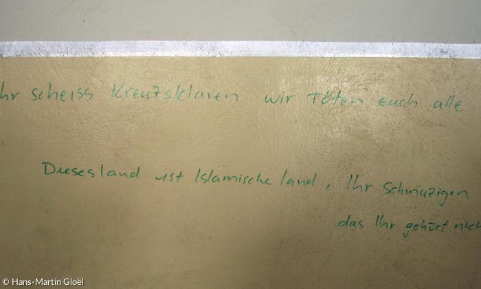 Graffito eines IS-Kämpfers mit Deutschkenntnissen in einer Seitenkapelle der Kirche von Batnaya: »Oh Ihr scheiss Kreuzsklaven wir töten euch alle. Dieses land ist Islamische land, Ihr Schmuzigen, das Ihr gehört nicht da hier«.