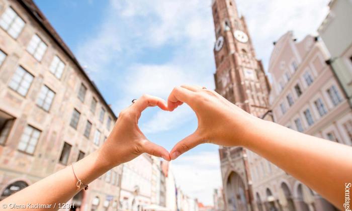 Hände bilden ein Herz vor dem Kirchturm der Landshuter Stadtpfarrkirche.