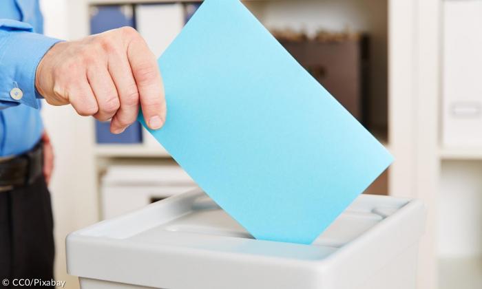 Ein blauer Zettel wird in eine Wahlurne gesteckt