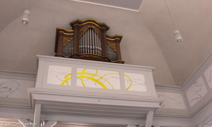 Der innenraum der Evangelischen Kirche Weisendorf. Man sieht zwei Stöcke mit einer weißen Empore. Auf der Empore sind gold- und silberfarbene Wellen. Auf der Empore im zweiten Stock befindet sich eine braun-silberne Orgel.