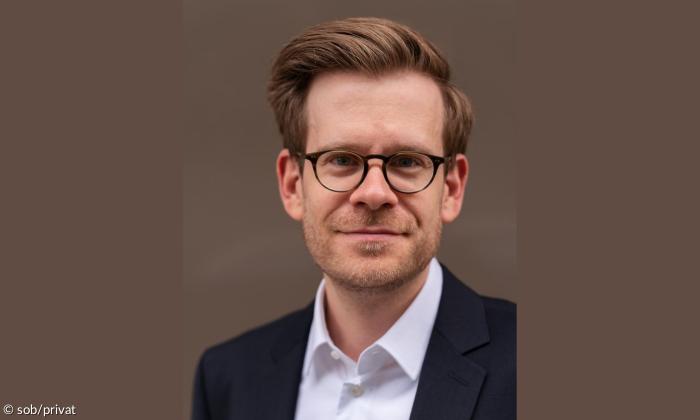 Ein Porträt von Andreas Braveny. Er trägt einen Anzug und eine Brille, lächelt in die Kamera und hat braune Haare.