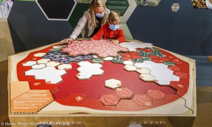 Eine Frau und ein Kind stehen vor einer riesigen Pizza aus Pappkarton. Die Zutaten sind als 6-Ecke vorhanden und können auf die Pizza gelegt werden.