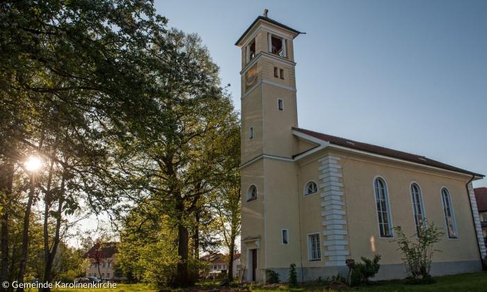 Die Hinterseite der Karolinenkirche im Sonnenschein. Zu sehen sind einige Fenster und der Kirchturm neben einem großen Baum, durch den das Sonnenlicht scheint.