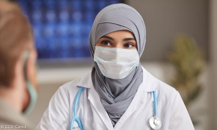 Eine Frau, die eine Maske und einen Arztkittel trägt. Um ihren Hals liegt ein Stethoskop. Sie trägt ein graues Kopftuch und schaut ihren Gegenüber an, der nur verschwommen sichtbar ist.
