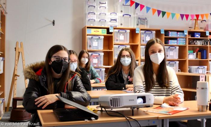 Kinder mit Masken sitzen an drei Tischen und blicken in die Kamera.