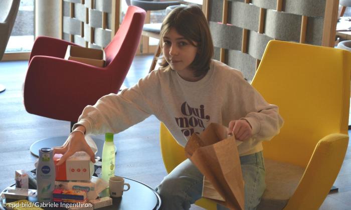 Ein Mädchen mit kinnlangen Haaren, das verschiedene Lebensmittel in eine Tüte packt. Sie trägt einen langärmligen Pullover mit der Beschrifung "oui, mon cheri" und blickt direkt in die Kamera.