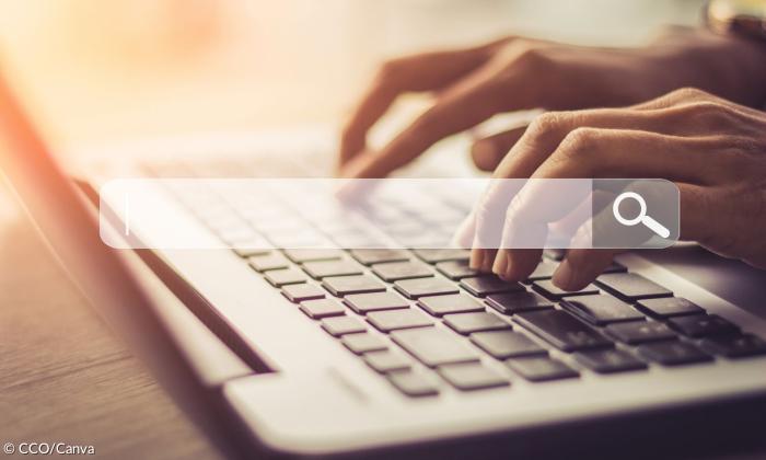Eine Person sitzt an einem Laptop. Man sieht nur die Hände. im Vordergrund ist eine Suchzeile zu sehen.