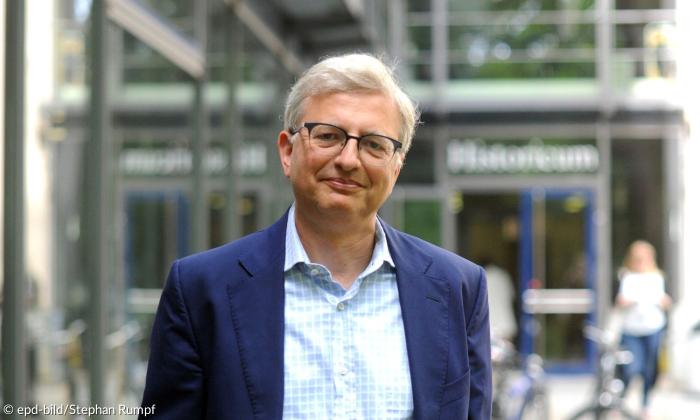 Ein weißhaariger weißer Mann im blauen Sakko steht vor einem Gebäude