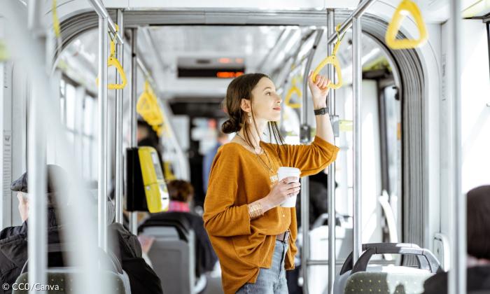 Eine junge Frau steht in einer Bahn und hält sich am Griff fest, in der anderen Hand ein Kaffee