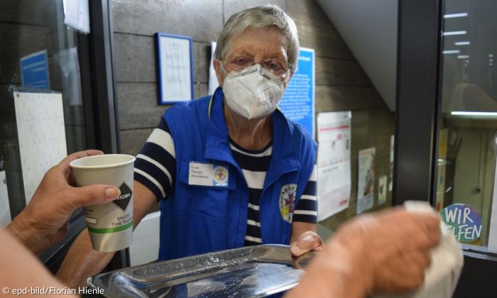 Eine Frau in blauer Weste und gestreiftem Kurzarmshirt reicht einem Menschen ein Tablett und einen Kaffee.