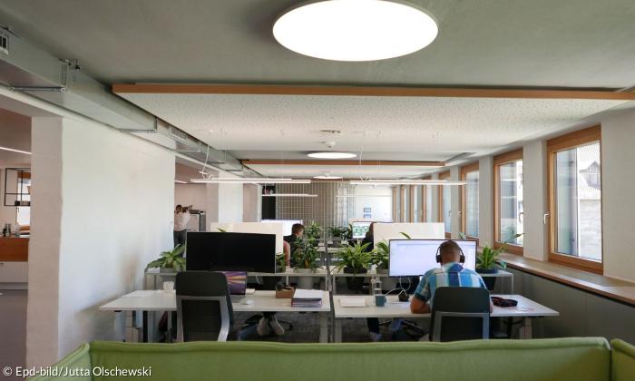 Blick in ein Büro mit Tischen, auf denen Pflanzen und Computer stehen. Vor einem Computer sitzt ein Mann in einem karierten Hemd, der Kopfhörer trägt.