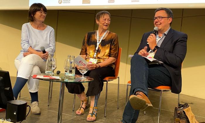 Ein Foto vom Panel "So kann gendern im Radio und TV gelingen" auf dem Podium sitzen Links Christibe Olderdissen, in der Mitte Moderatorin Elke Zimmermann und rechts Thomas Apfel, der gerade ein Mikrophon in der Hand hält.