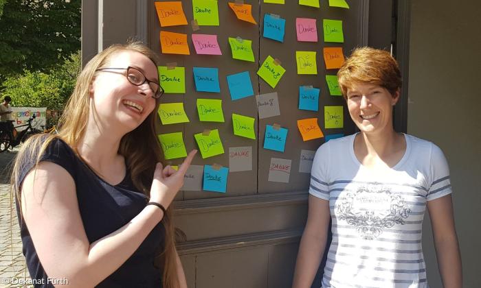 Zwei Frauen vor einer Vitrine. In der Virtrine sind bunte Post-Its auf denen Danke steht. Die Frau links ist ca Ende zwanzig, hat lang blonde Haare, eine Brille auf und trägt ein schwarzes Tshirt. Die Frau rechts hat kurze brauene Haare, ist ca Mitte dreisig und trägt ein weißes Shirt mit blauen Streifen drauf