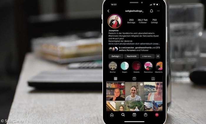 Ein Smartphone an ein Wasserglas gelehnt. Es steht auf einem Schreibtisch. Auf dem Handy ist das Instagramprofil von Seligkeitsdinge zu sehen.