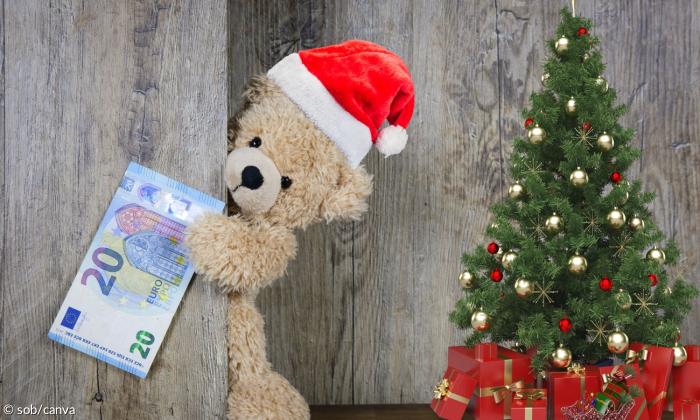 Ein Teddybär schaut aus einer Türe raus und hält einen 20 Euro Schein. Er trägt eine Weihnachtsmütze. Rechts von ihm steht ein kleiner Baum, der mit roten und goldenen Kugeln geschmückt ist. Darunter liegen Geschenke, die in rotes Geschenkpapier eingepackt sind.