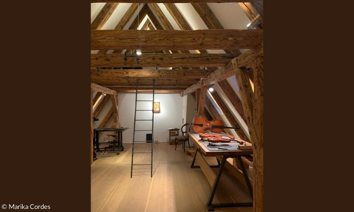 Dachgeschoss mit Holzbalken, Geigen und ein Cello rechts auf einem Tisch, eine Eisenleiter führt in den Dachspitz