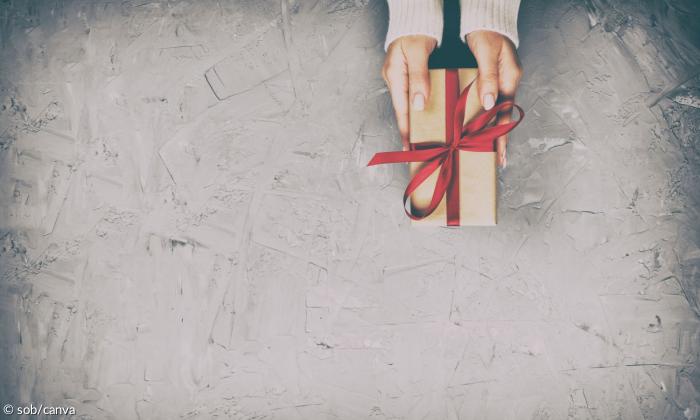 ein grauer Hintergrund. Von oben ragen ein paar weibliche Hände ins Bild, die ein Geschenk halten. Das Geschenk ist in braunes packpapiert verpackt und hat eine rote Schleife.