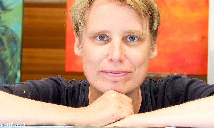 Porträt von einer Frau mit kurzen blonden Haaren. Sie schaut direkt in die Kamera und stützt ihr Kinn auf ihre Faus. Die Arme liegen auf einem Tisch auf. Im Hintergrund hängt ein orangenes Bild