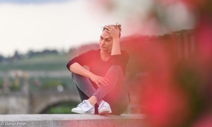 Eine junge Frau sitzt auf einer Mauer. Ihre Beine sind vor ihrem Körper über Kreuz aufgestellt. Mit ihrer linken Hand hält sie ihren Kopf.