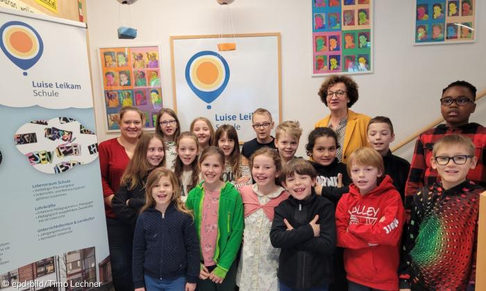 Eine Gruppe Grundschüler steht neben einem Banner mit der Aufschrift Luise Leikam Scvhule. Hinter den Kindern steht eine ältere Frau und an der Wand hängen Bilderrahmen mit bunten Popart Fotos von Luise Leikam.