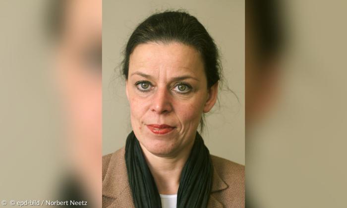 Porträt einer Frau mit dunklen Haaren, die sie nach hinten gebunden hat. Sie trägt hellroten Lippenstift, einen beigenen Blazer und einen schwarzen Schal