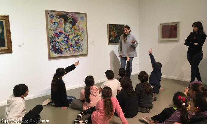 Vor mehreren an einer Wand hängenden Gemälden Gemälden sitzen kleine Kinder und schauen aufmerksam einer Frau zu, die erklären vor einem Bild steht.