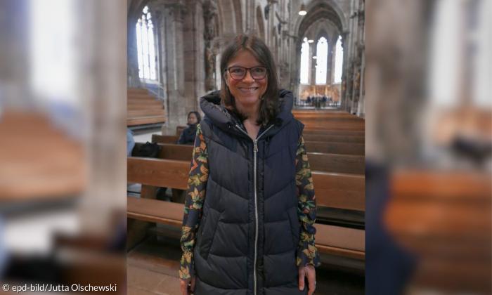 Eine Frau Mitte Vierzig blickt mit einem Lächeln in die Kamera. Sie steht vor einer Kirchenbank in einer großen Kirche. Im Hintergrund sieht man viele Reihen von Kirchenbänken, auf denen vereinzelt Menschen sitzen.