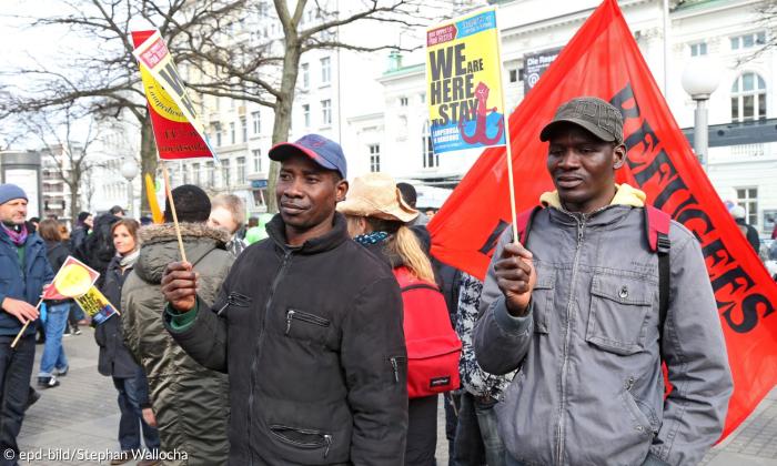 Zwei Männer mit Fahnen und Schildern, dahinter mehr Demonstrant*innen