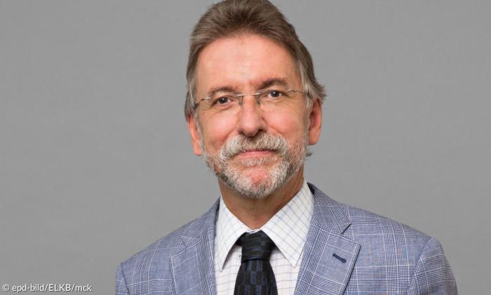 Mann Ende vierzig blickt mit leicht angewinkeltem Mund frontal in die Kamera. Man sieht im Porträt sein Gesicht und den Schulterbereich. Er trägt einen grauen Anzug und Krawatte sowie eine Brille.