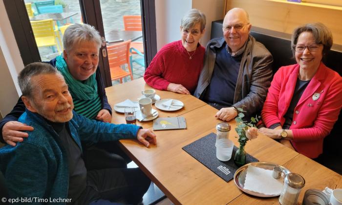Zwei Paare, die sich in den Armen halten sitzen mit einer weiteren Frau an einem Café-Tisch. Sie lächeln alle in die Kamera und sind fünfzig bis siebzig Jahre alt.