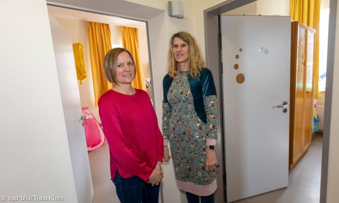 zwei Frauen Mitte vierzig blicken in die Kamera. Sie stehen auf einem gang und jeweils zur linken Seite und rechten Seite sieht man zwei offene Zimmetüren.