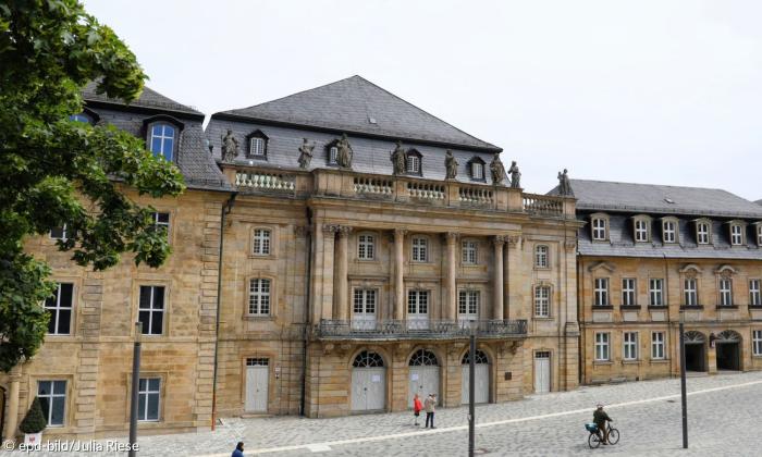 Man sieht ein großes Opernhaus, das das gesamt Bild für sich beansprucht. Davor sind vereinzelnd Menschen auf der Straße zu sehen. 