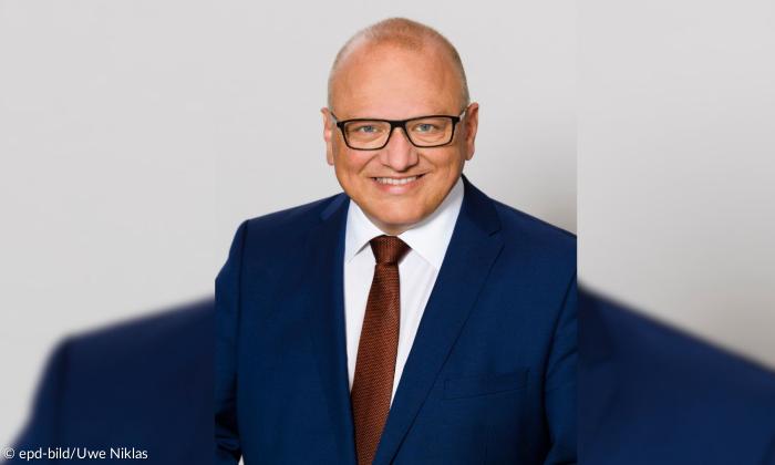 Porträtaufnahme eines Mannes, der frontal in die Kamera blickt. Er hat eine Glatze, trägt eine Brille sowie einen dunkelblauen Anzug. 