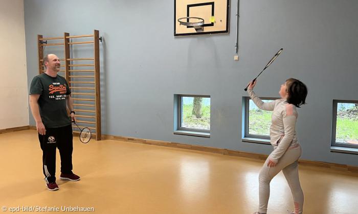 Simon Schneidereit spielt mit seiner elfjährigen Tochter Federball in einer kleinen Turnhalle mit kleinen Fenstern am Boden und einem tiefhängenden Basketballkorb an der Wand. 