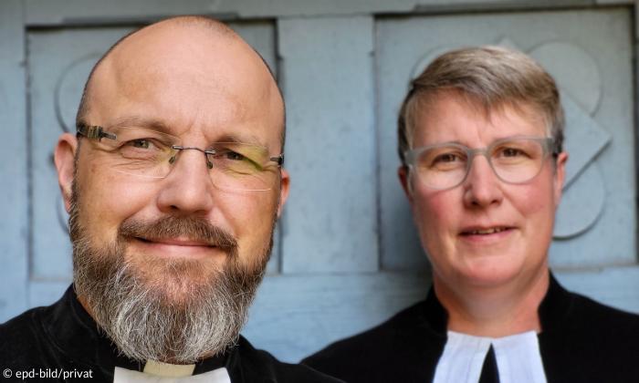 Ein Mann und eine Frau blicken frontal in die Kamera. Ihre  Man sieht nur ihren Kopf und ihren Schulterbereich. Sie wirken fröhlich, da sie die Mundwinkel leicht angewinkelt haben. Beide tragen einen Talar.