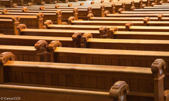Man sieht mehrere Reihen von Kirchenbänken, die sich vor dem Betrachter aufbauen. Sie sind alle leer.