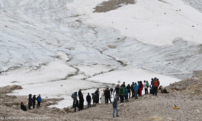 eine Menschentraube steht vor einem kleinen Gletscher. Zu dessen Fuß sieht man sehr viel Gestein.