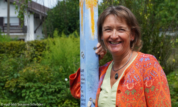 Eine Frau mit braunen halblangen Haaren lehnt in einem Garten an einem hellblau, gelb bemalten Pfahl.. Sie lächelt in die Kamera und trägt ein gelbes Oberteil, darüber eine orangene Jacke mit bunten Ornamenten.