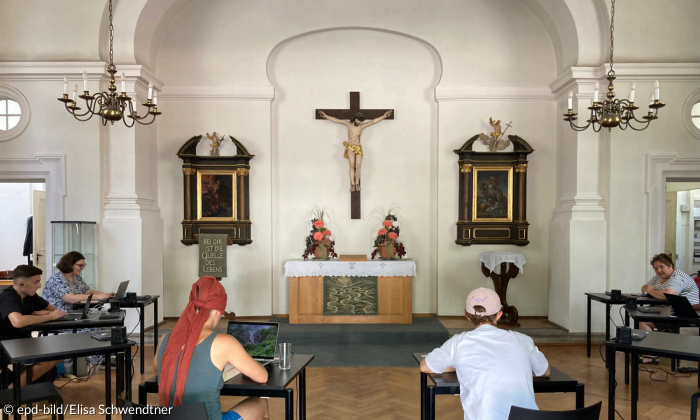 Sechs Studierende sitzen im Halbkreis and Schreibtischen vor einem Altar über de, ein kreuz hängt. Rechts und links hängt jeweils ein Kronleuchter.