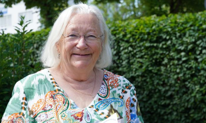 Porträtaufnahme einer Frau Mitte sechzig. Sie hat lange, weiße Haare und trägt eine Brille. Die Frau lach in die Kamera.
