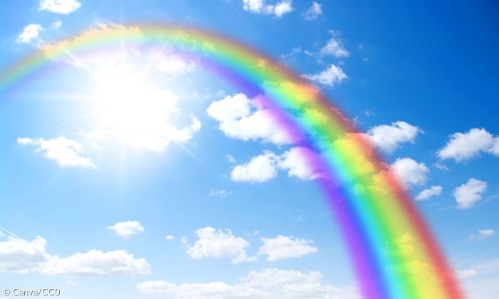 Ein Regenbogen erstreckt sich von der linken oberen Ecke zur rechten unteren Ecke über das ganze Bild. Hinter ihm sind die strahlende Sonne, Wolken und blauer Himmel zu erkennen. 