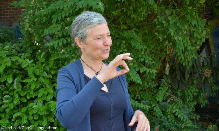 Seitenaufnahme einer Frau Mitte vierzig. Sie hat kurze graue Haare und gestikuliert mit ihrer rechten Hand, wobei Zeigefinger und Daumen ein Oval formen. 