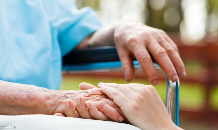 Man sieht die Hände einer älteren Person, die auf einem Stuhl sitzt. Eine Hand liegt auf einer Metallarmlehne und die Hand einer weiteren Person hält die andere Hand der sitzenden Person.