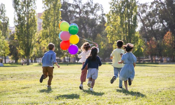 5 Kinder laufen über eine Wiese, eines hält bunte Luftballons an einer Schnur