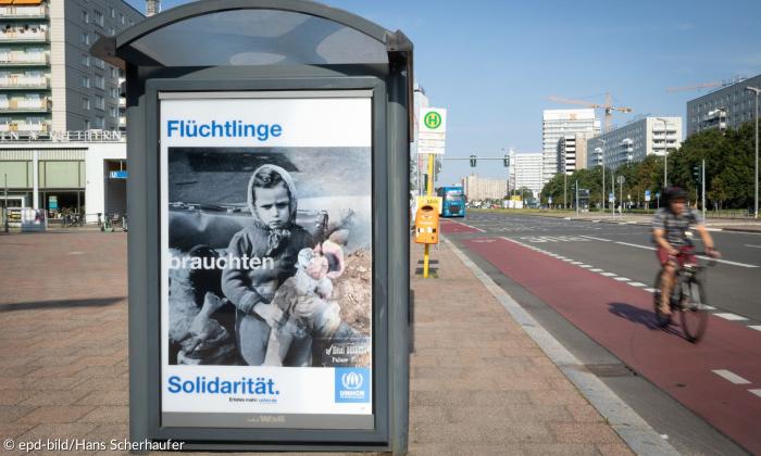 Bushaltestelle mit einem Plakat, auf dem "Flüchtlinge brauchten Solidarität" steht, dazu ein schwarzweiß Bild eines vertriebenen Mädchens