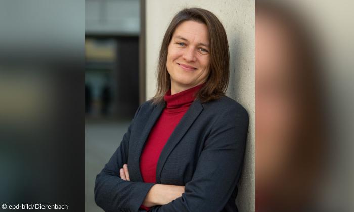 Frau mit braunen Haaren steht an einer Wand und lächelt