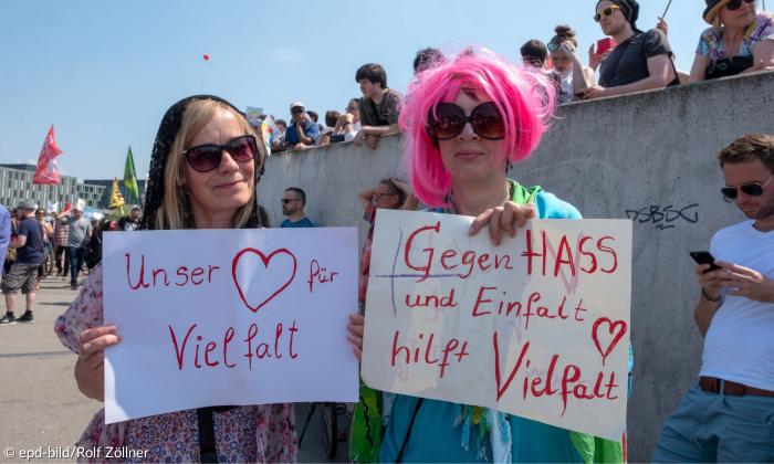 Zwei Personen auf einer Demo, auf ihren Schildern steht "Unser Herz für Vielfalt" und "Gegen Hass und Einfalt hilft Vielfalt"