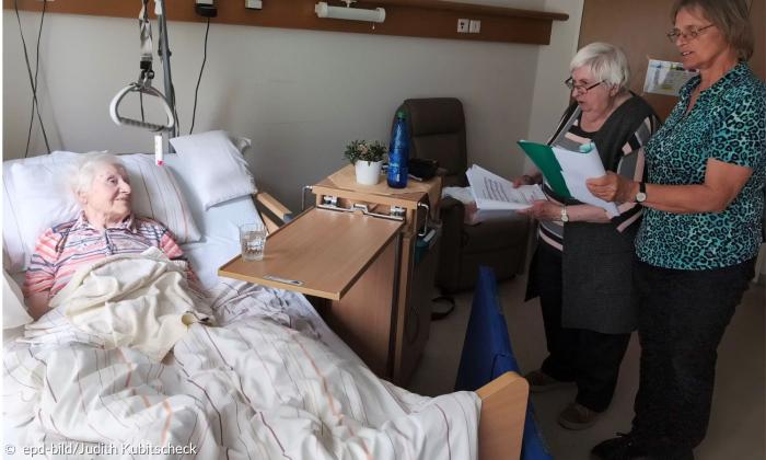 Zwei Frauen singen am Bett einer liegenden Seniorin