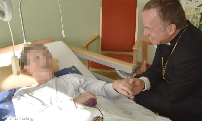 Ein Mann sitzt am Bett einer Frau, deren Gesicht verpixelt ist